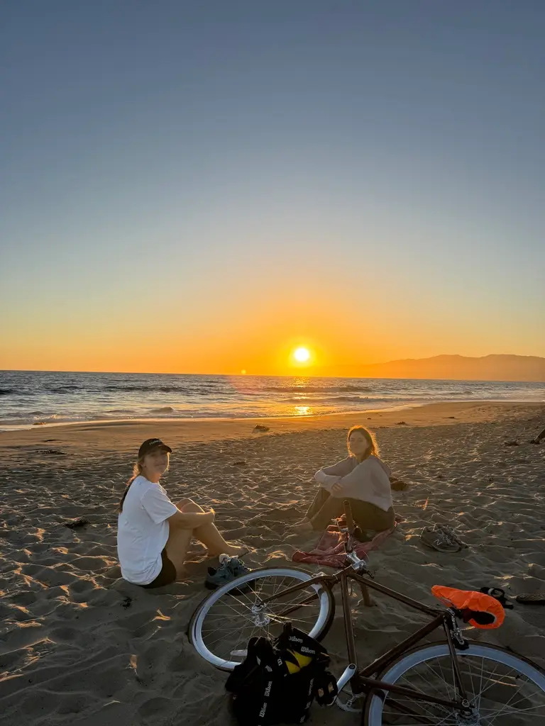 Two riders sitting beside a bike on the sand at sunset on the Los Angeles coast