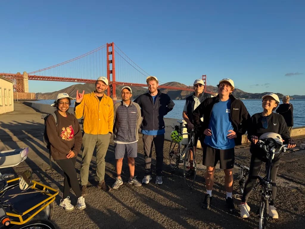 Seven riders in matching caps with their bikes in front of the Golden Gate Bridge at sunrise