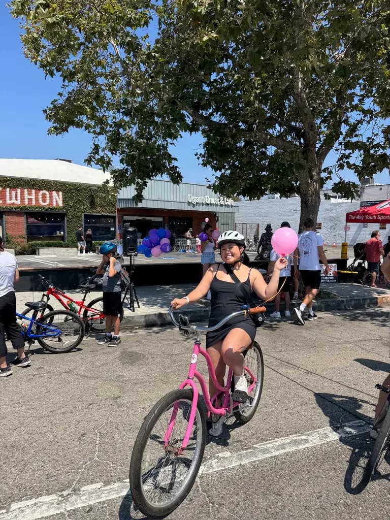 A rider in a helmet on a pink cruiser bike, holding a pink balloon on a car-free street in Los Angeles